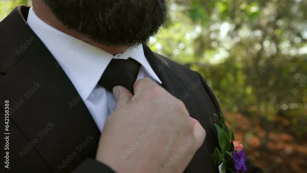 Strong male hand of groom with thick black beard, fixing black tie on neck. Silhouette of groom in white shirt, black jacket, black tie, standing outdoors. High quality 4k footage