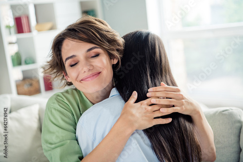 Photo of dreamy adorable young girls wear shirts sitting sofa embracing closed eyes indoors apartment room