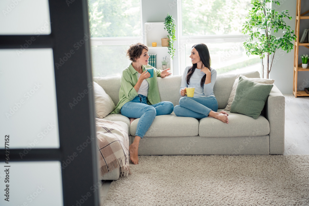 Full size photo of two positive lovely girls sit comfy couch drink coffee communicate enjoy free time flat indoors