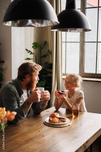 Smiling child holding donut and talking to tattooed father with coffee during breakfast in kitchen 
