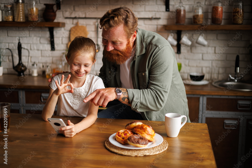 Tattooed father and freckled kid gesturing and using smartphone together near sweet pastry in kitchen in morning