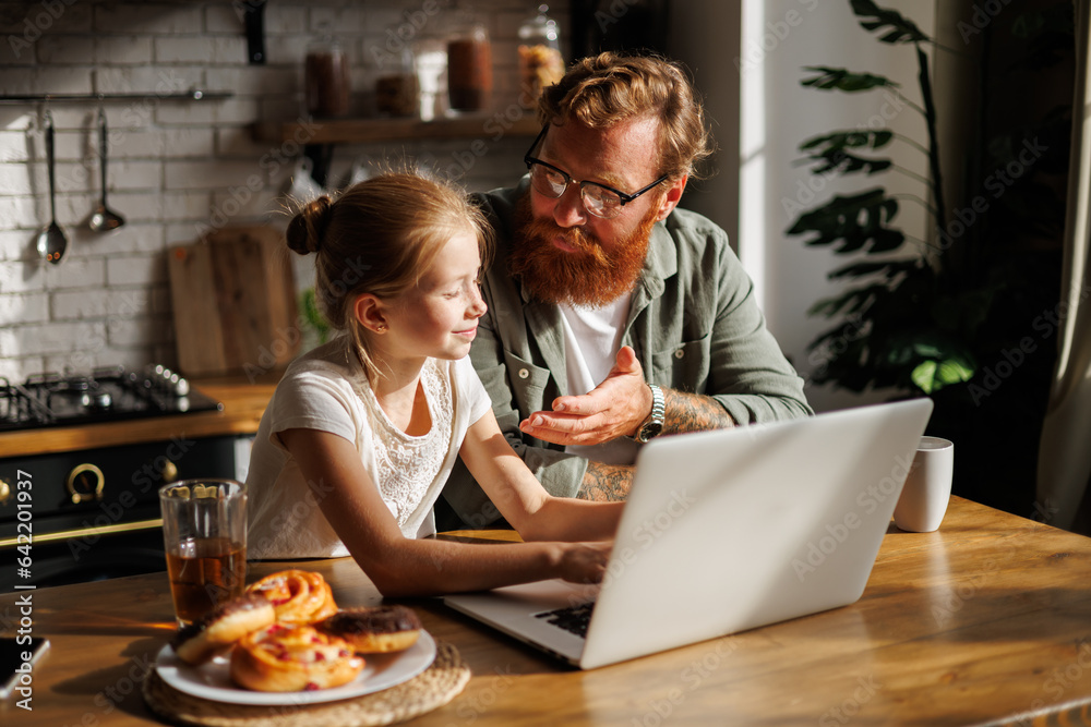 Tattooed and bearded dad talking to smiling daughter using laptop near breakfast in morning in kitchen  - obrazy, fototapety, plakaty 