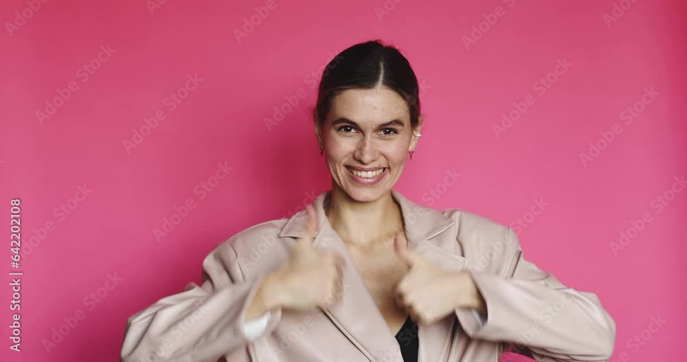 Optimistic brunette woman makes thumbs up gives approval shows excellent gesture says nice job gives positive feedback shares good opinion dressed in leather jacket isolated over pink background. Optimistic brunette woman makes thumbs up gives approval shows excellent gesture says nice job gives positive feedback shares good opinion dressed in leather jacket isolated over pink background.