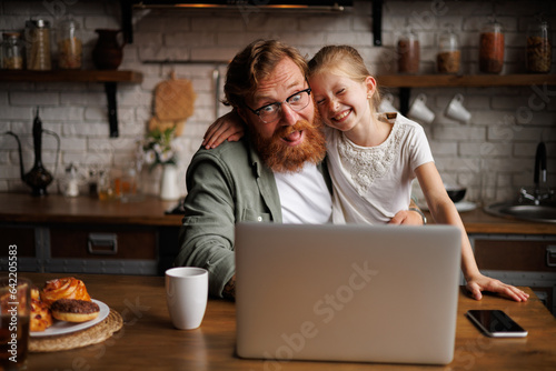 Bearded man in eyeglasses having fun with cheerful daughter near laptop and pastry in kitchen 