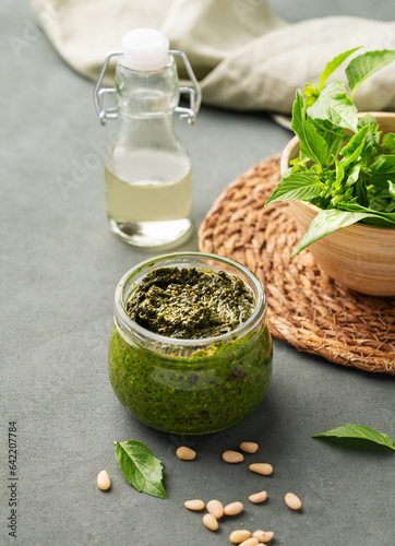 Traditional Italian pesto in a jar with green basil, pine nuts, cheese and olive oil on a green background. A classic sauce for spaghetti or bruschetta.