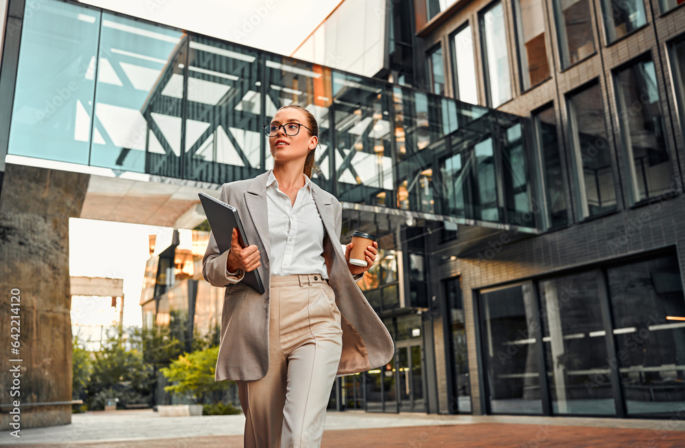 Confident beautiful young stylish modern business woman in jacket and glasses holding laptop and coffee looking to the side and going to a meeting at work. Rushing to work.
