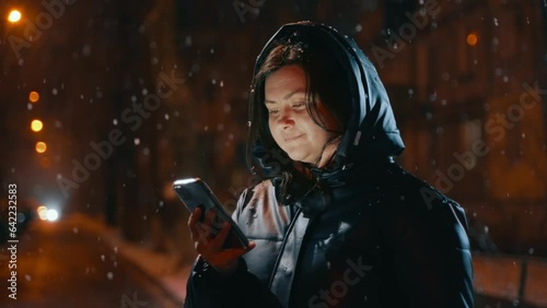 Woman outdoors in a hood with a phone in the bokeh of blurry light, it's snowing