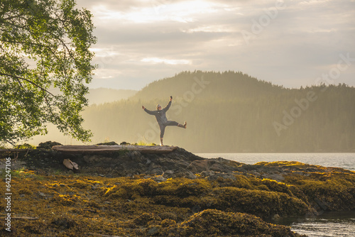 happy in nature: a man in silhouette throws up his arms and a leg in joy standing aon a rocky shore with a forest covered mountain and sea behind him backlit room for text