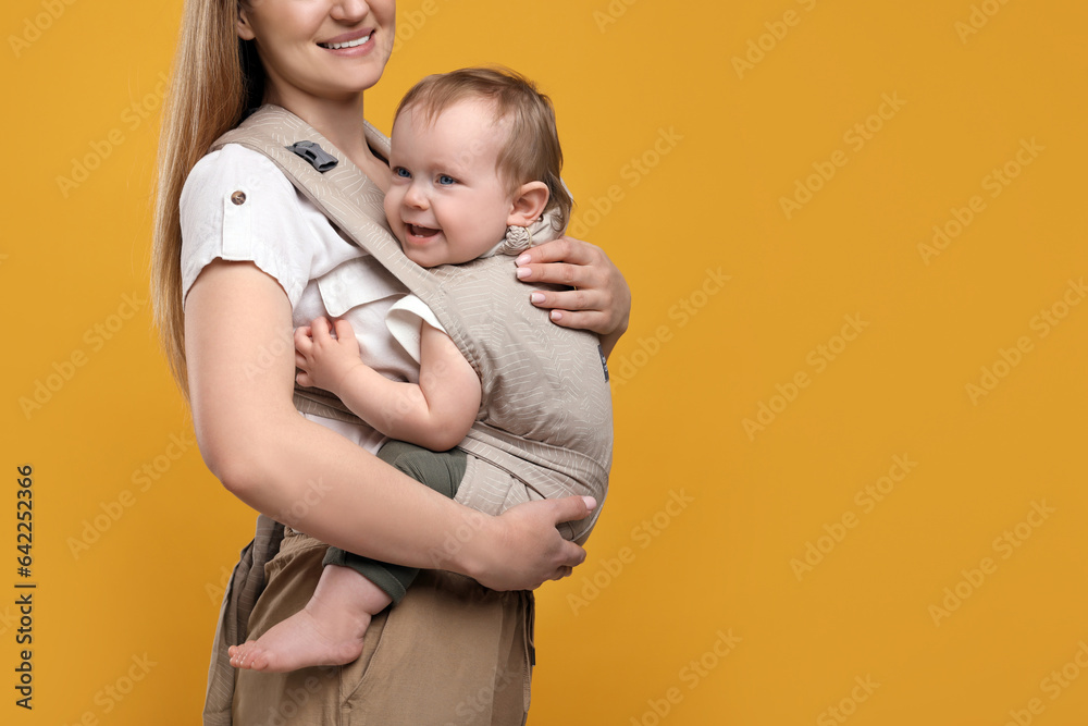 Mother holding her child in sling (baby carrier) on orange background, closeup. Space for text