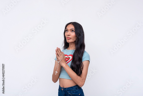 A religious asian trans woman looking up an praying with a rosary. Isolated on a white background.
