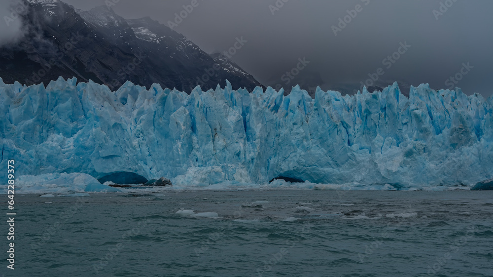 Fragment of the Perito Moreno glacier. A wall of cracked blue ice with prickly spikes rises above a turquoise lake. Ice floes float in the water. Mountains in fog and clouds. Argentina. Fragment of the Perito Moreno glacier. A wall of cracked blue ice with prickly spikes rises above a turquoise lake. Ice floes float in the water. Mountains in fog and clouds. Argentina.