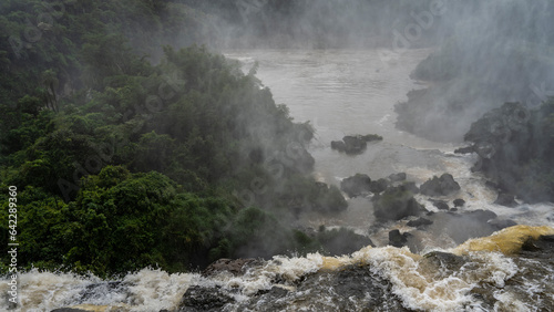 A stream of seething water collapses from a ledge. Spray and mist in the air. Boulders in the riverbed. Tropical vegetation around. Iguazu Falls. Argentina.