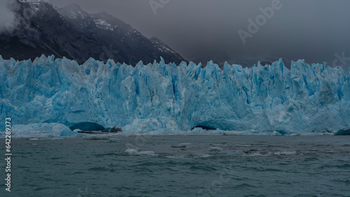 Fragment of the Perito Moreno glacier. A wall of cracked blue ice with prickly spikes rises above a turquoise lake. Ice floes float in the water. Mountains in fog and clouds. Argentina.