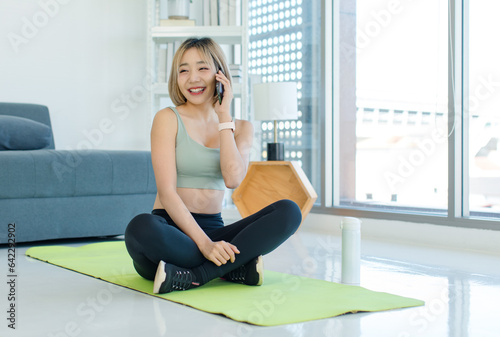 Free Download Asian young female athlete teenager in sportswear sport bra sitting smiling on yoga pilates mat holding using smartphone in hand in gym after training exercising working out. - GFXLoad