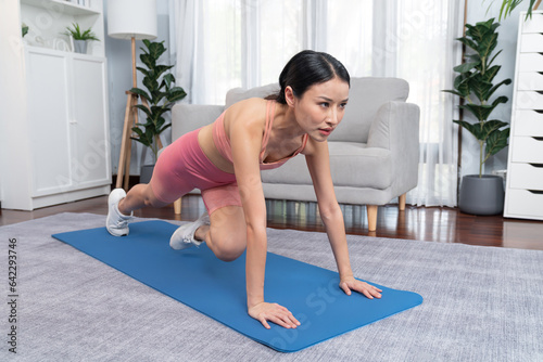 Asian woman in sportswear doing burpee on exercising mat as home workout training routine. Attractive girl engage in her pursuit of healthy lifestyle and fit body physique. Vigorous photo