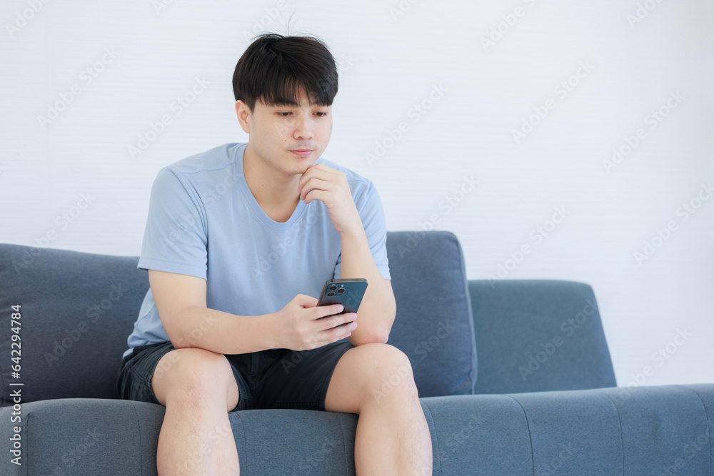 Asian young handsome fit male athlete teenager in sportswear sneakers sitting on cozy sofa couch on call with smartphone surfing browsing internet online at home. Asian young handsome fit male athlete teenager in sportswear sneakers sitting on cozy sofa couch on call with smartphone surfing browsing internet online at home.