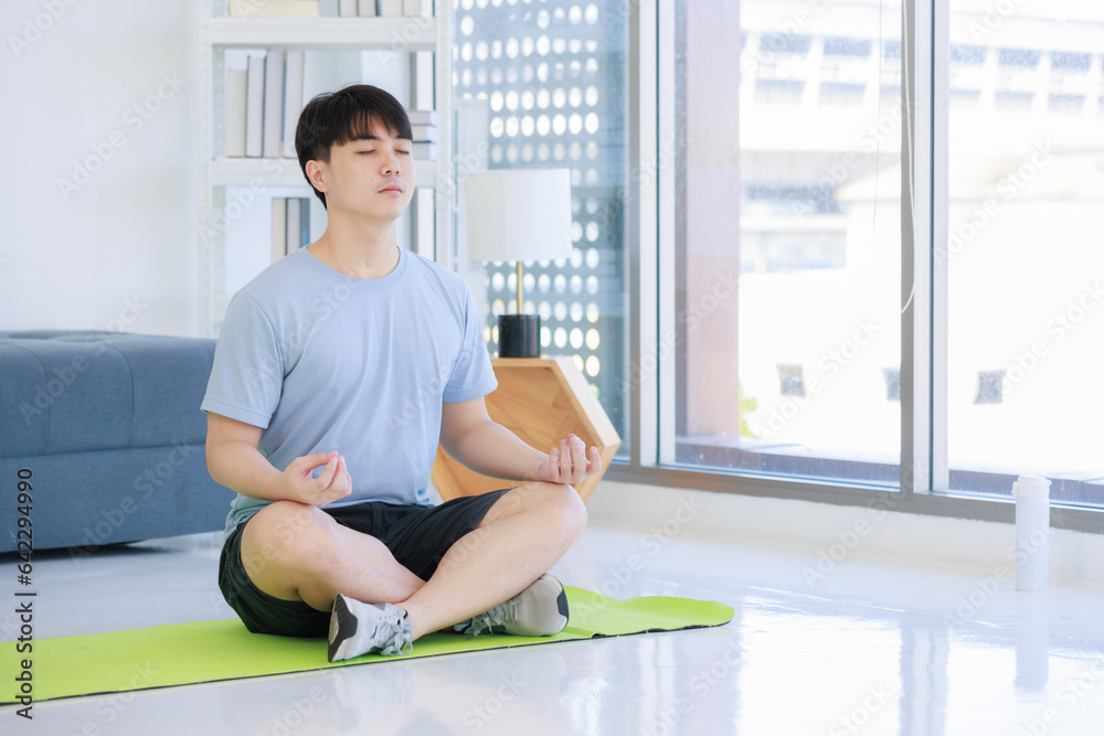 Asian young handsome fit male athlete teenager in sportswear sneakers sitting closed eyes meditating on yoga mat with lotus posture exercising training together in living room at home. Asian young handsome fit male athlete teenager in sportswear sneakers sitting closed eyes meditating on yoga mat with lotus posture exercising training together in living room at home.