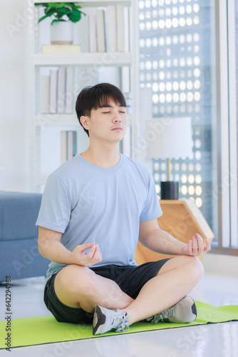 Asian young handsome fit male athlete teenager in sportswear sneakers sitting closed eyes meditating on yoga mat with lotus posture exercising training together in living room at home.
