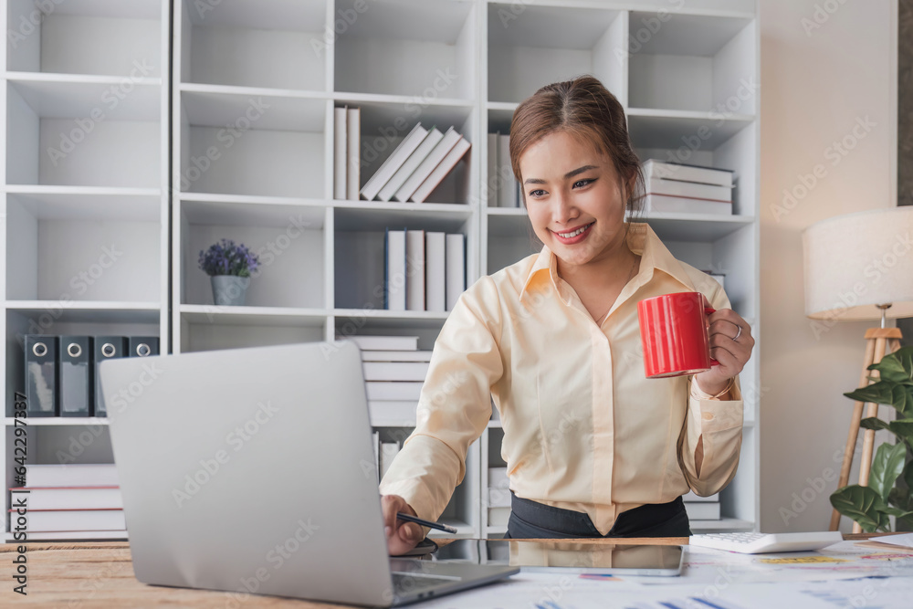 Beautiful and happy young Asian businesswoman looking at her laptop screen while enjoying her morning coffee at her desk in the office.