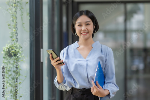 Portrait of a smiling asian businesswoman using mobile phone and holding document in the office. © amnaj