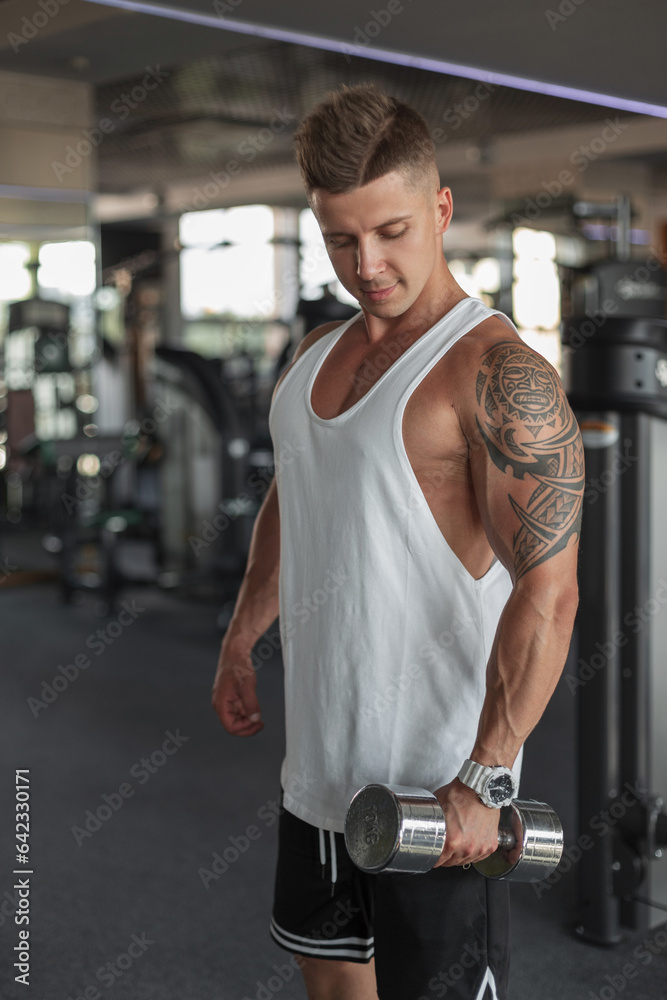 Cool stylish muscular athletic man with a haircut in a white tank top with dumbbells in the sport gym Cool stylish muscular athletic man with a haircut in a white tank top with dumbbells in the sport gym