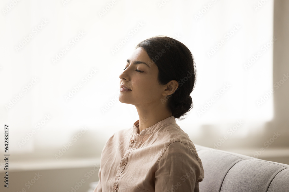 Calm tranquil young Indian woman meditating at home, sitting on sofa with straight back and closed eyes, breathing fresh air, doing yoga breathwork, practicing relaxation, stress relief, meditation Calm tranquil young Indian woman meditating at home, sitting on sofa with straight back and closed eyes, breathing fresh air, doing yoga breathwork, practicing relaxation, stress relief, meditation