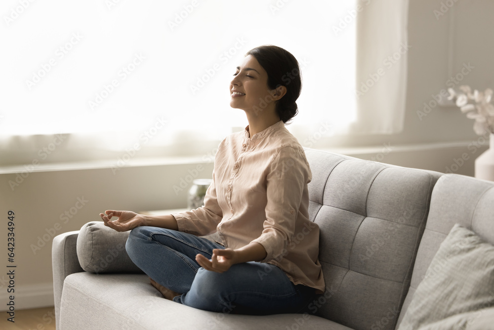 Happy peaceful young Indian yogi woman meditating on home couch, keeping lotus yoga pose, zen mudra, calm hand gesture, smiling with closed eyes, taking deep breath of fresh air Happy peaceful young Indian yogi woman meditating on home couch, keeping lotus yoga pose, zen mudra, calm hand gesture, smiling with closed eyes, taking deep breath of fresh air