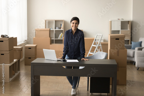 Happy successful beautiful Indian entrepreneur girl standing at home workplace with laptop, looking at camera, smiling. Small business project, internet store owner full length portrait © fizkes