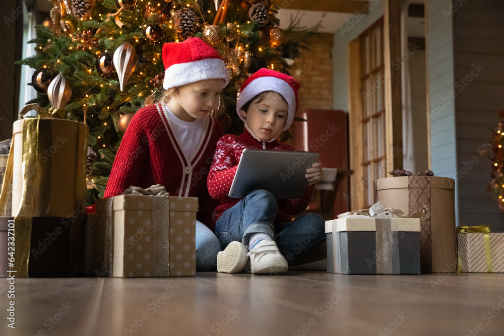 Happy interested little children boy girl in Santa Claus hats sitting on warm heated floor near decorated Christmas trees and gifts, playing online games together on digital tablet, tech addiction. Happy interested little children boy girl in Santa Claus hats sitting on warm heated floor near decorated Christmas trees and gifts, playing online games together on digital tablet, tech addiction.