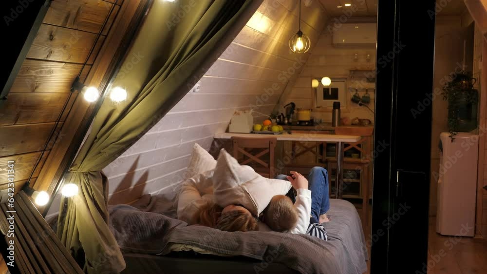 Mother and son have fun lying on the bed in a small wooden triangular house, they throw pillows up. Vacation in a country house in autumn. View through a window with flashlights and a thick curtain. Mother and son have fun lying on the bed in a small wooden triangular house, they throw pillows up. Vacation in a country house in autumn. View through a window with flashlights and a thick curtain.