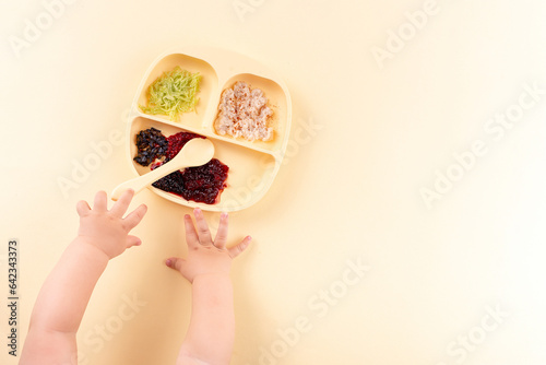Children's silicone plate with food, the baby's hands reach for dinner. Feeding on a yellow background.