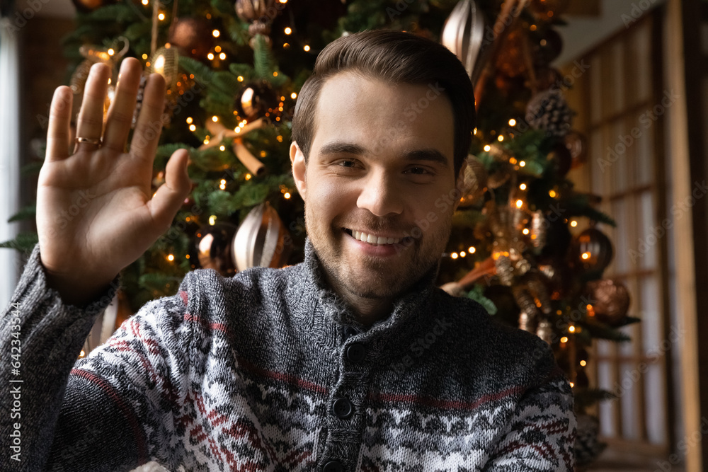 Happy young handsome man waving hand starting web camera video call distant conversation with decorated Christmas tree on background, congratulating friends or family parents with New year holidays. Happy young handsome man waving hand starting web camera video call distant conversation with decorated Christmas tree on background, congratulating friends or family parents with New year holidays.