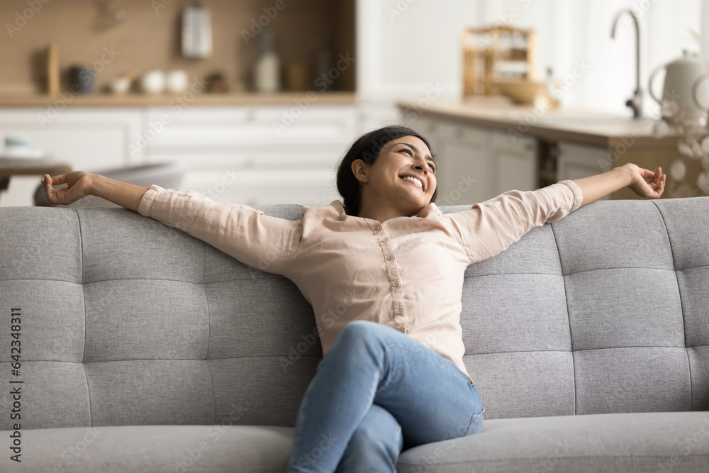 Cheerful carefree beautiful Indian woman relaxing on home sofa, stretching body on soft back with open arms, breathing fresh pure air, enjoying domestic comfort, happiness, looking away, daydreaming Cheerful carefree beautiful Indian woman relaxing on home sofa, stretching body on soft back with open arms, breathing fresh pure air, enjoying domestic comfort, happiness, looking away, daydreaming