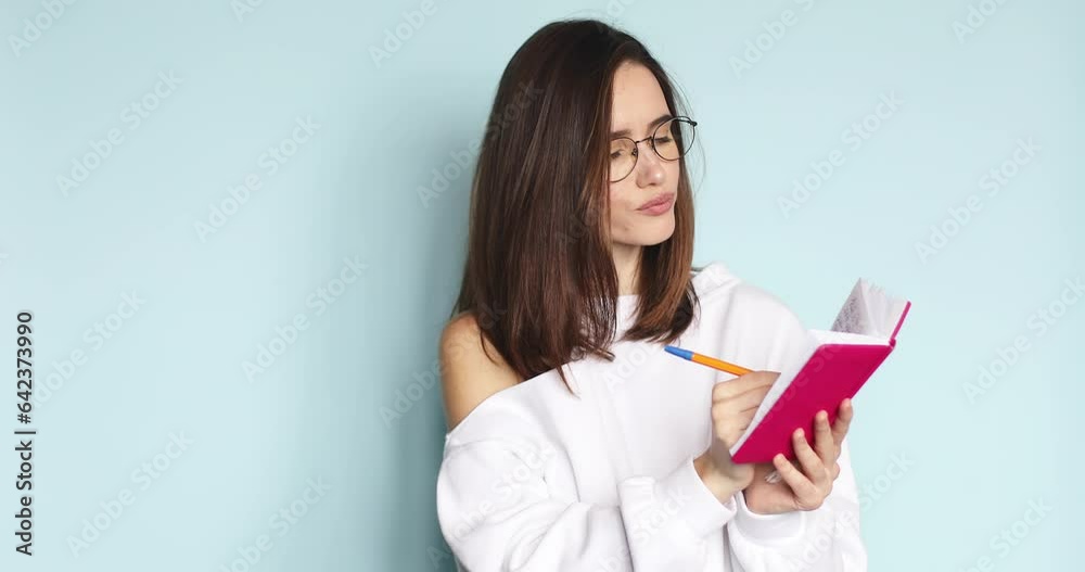 Young beautiful girl writing in the diary her romantic story, poem while she is smiling, dreaming, thinking. Creative woman in eyeglasses writing with pen at her pink notebook on blue background. Young beautiful girl writing in the diary her romantic story, poem while she is smiling, dreaming, thinking. Creative woman in eyeglasses writing with pen at her pink notebook on blue background.