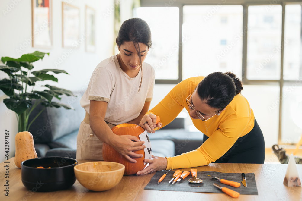 Young ethnic women standing at table with Halloween paper print on pumpkin Young ethnic women standing at table with Halloween paper print on pumpkin