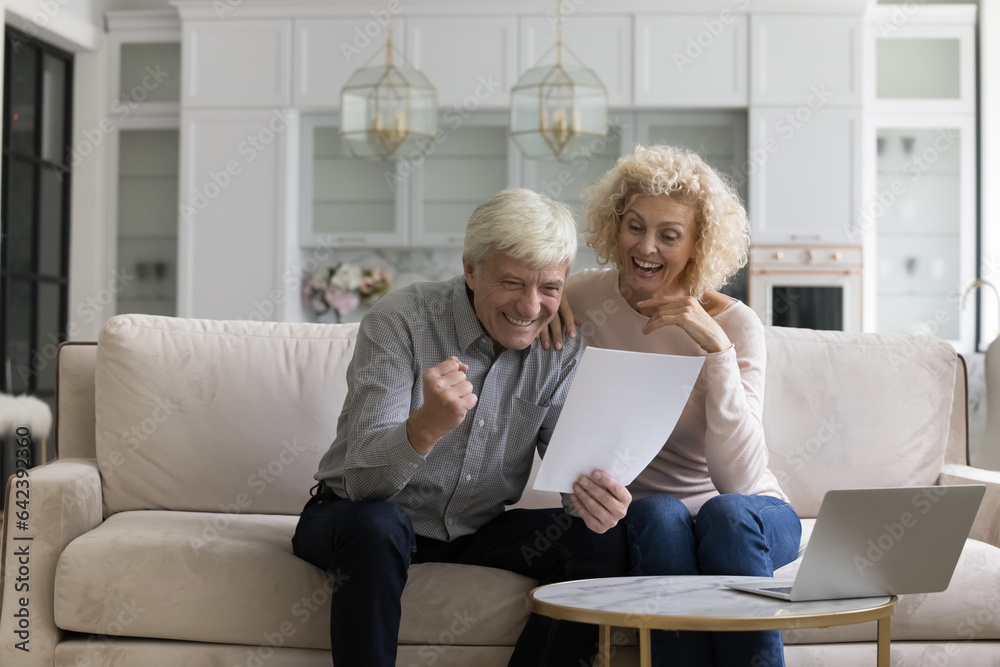 Cheerful excited old mature couple reading document with winner yes hand, celebrating financial success, achieve, investment income, good surprising news, laughing, shouting for joy