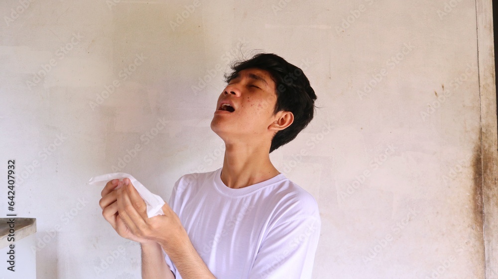 an asian man in a white t-shirt has a cold and uses a tissue to cover his sneeze an asian man in a white t-shirt has a cold and uses a tissue to cover his sneeze