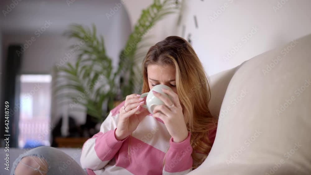 Young beautiful Caucasian woman with blond hair drinking from cup of tea or coffee in living room. Calm single female sitting on sofa at home. Casual people with relaxed expression enjoying hot drink. Young beautiful Caucasian woman with blond hair drinking from cup of tea or coffee in living room. Calm single female sitting on sofa at home. Casual people with relaxed expression enjoying hot drink.