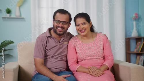 Happy smiling Indian middle aged couple posing or looking to camera at home on sofa - concept of married relationship, empty nesters and Quality Time. photo