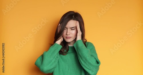 Woman with pain in her temples. Caucasian woman in green sweater suffering from stress or headache grimacing in pain, frowning from pain suffering unbearable migraine on yellow background. Medical.