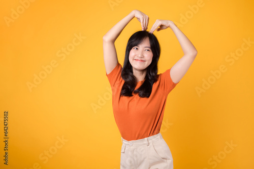 Warmth and authenticity shine through as a cheerful Asian woman in her 30s embraces love with a heart-shaped gesture and an infectious smile.