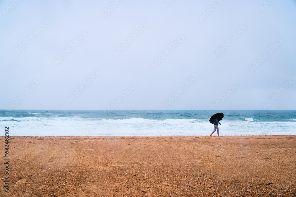 young adult with umbrella walking on the beach under the rain with rough sea
