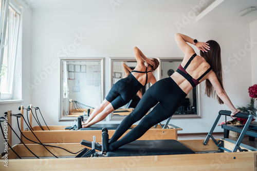 Fotografie, Obraz Two smiling fit-shaped females doing side plank with raised arm static core muscles exercise using pilates reformer machine in the sport athletic gym with a mirror wall