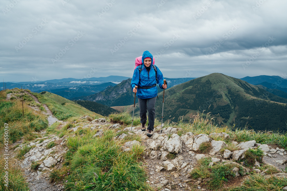 Obraz premium Young smiling hiker woman dressed rain suit walking by rocky Slovakian Mala Fatra mountain range using tracking poles with backpack. Active lifestyle concept image