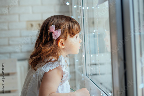 A child looks out the window waiting for spring. a girl in a white T-shirt, with a pink hairpin and brown hair. A child looks out the window waiting for spring
