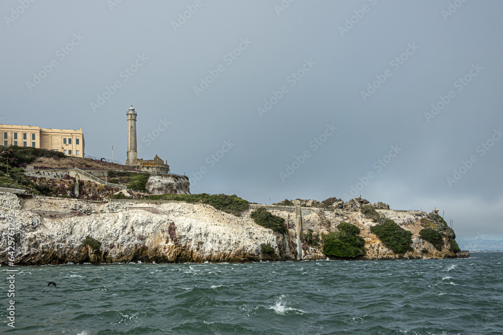 San Francisco, CA, USA - July 12, 2023: Alcatraz Island SW tip closeup with lighthouse. White guano covered rocky cliffs with sparse green bushes under light blue sky and darker greeenish bay water