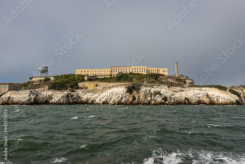 San Francisco, CA, USA - July 12, 2023: Alcatraz Island west rocky shite-guano covered cliffs. Beige Prison building and water tower on the left. Gray foggy sky and greenish bay water