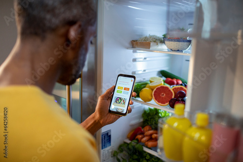 Middle aged african american man using a dieting app while looking through his fridge in the kitchen at home photo