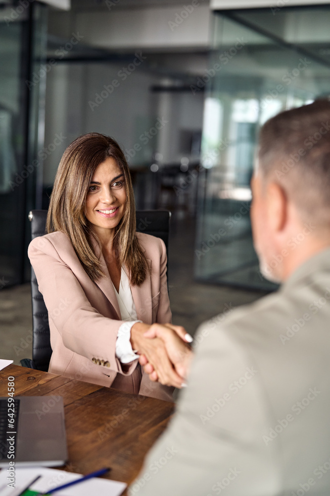 Happy mature Latin business woman manager lawyer handshaking client at office meeting. Smiling professional businesswoman and businessman shake having partnership agreement with handshake. Vertical
