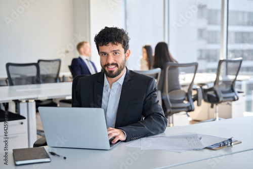 Smiling Latin young business man employee sitting at work with laptop. Confident happy office worker executive manager looking at camera at workplace in modern coworking or meeting room, portrait.